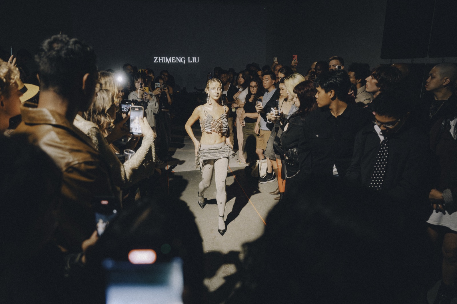 Seen from inside the crowd, a model walks the open floor beneath a designer's name on the back wall.