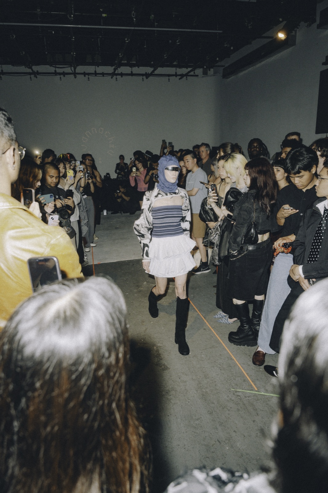 A model in a white skirt and tall black boots walks a runway that opens up through the middle of the crowd.
