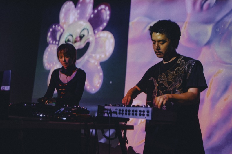 Two performers at a DJ and VJ table in front of a giant projected cartoon flower.