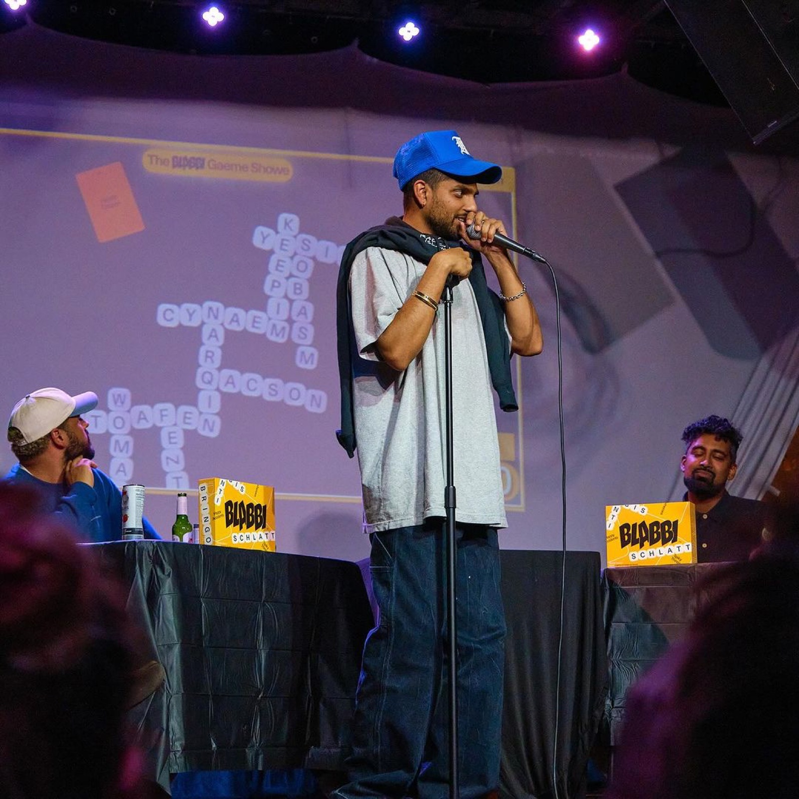 A comedian pitches into a microphone on a purple-lit stage while the projected game board glows behind him.