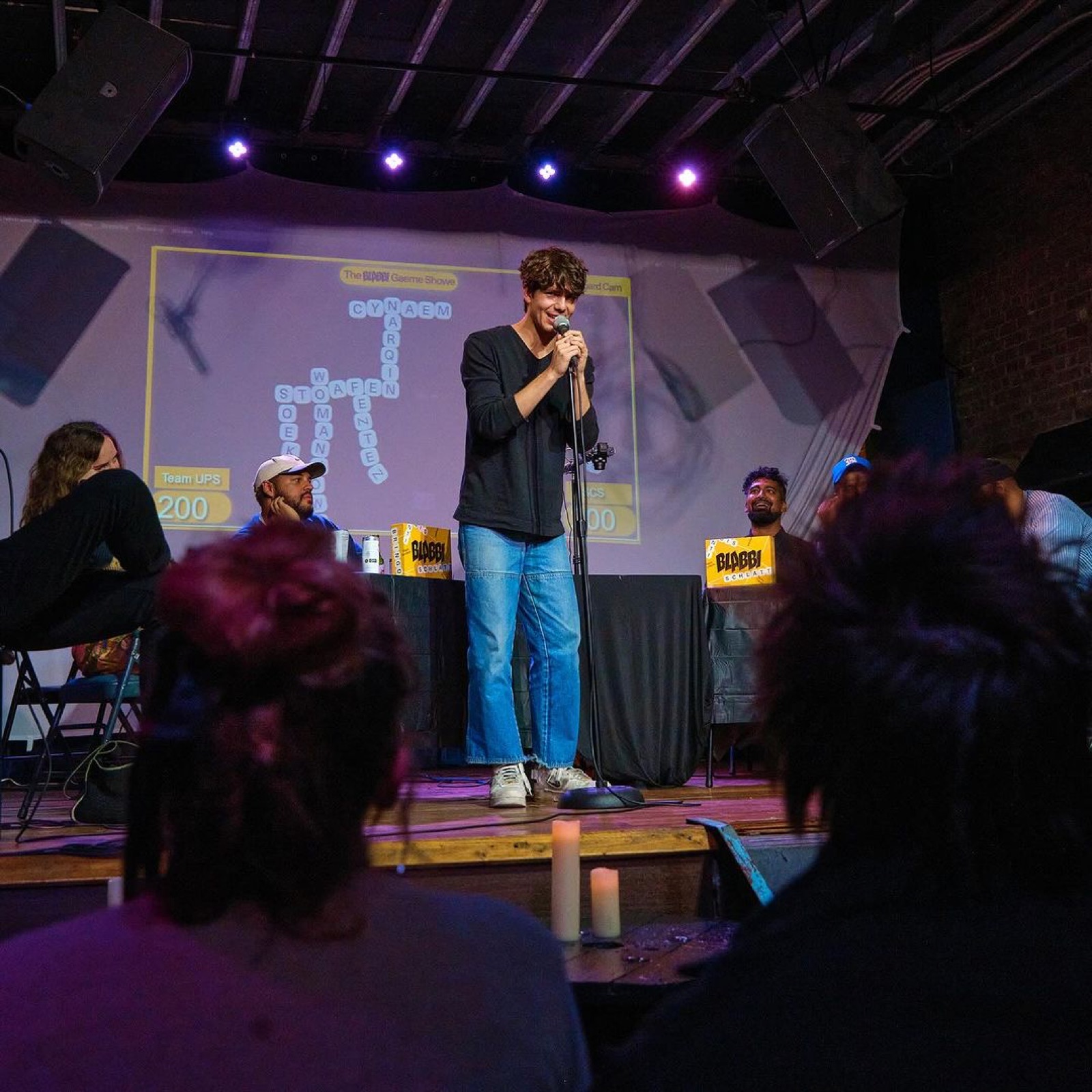A contestant in a white T-shirt pitches to the room with the projected tile board behind him.