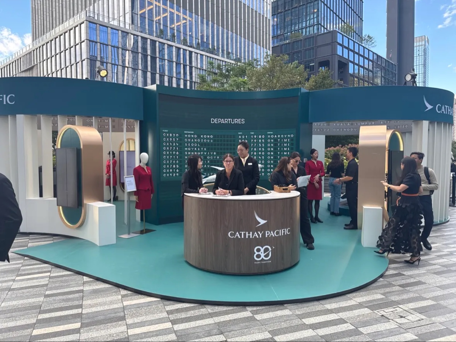 A check-in desk in front of a big green LED wall showing a departures board, at the Cathay Pacific pop-up.