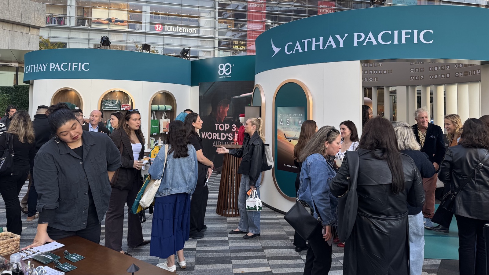 Outside the pop-up, a crowd gathers by curved teal walls with Cathay Pacific signage and an arched LED screen.