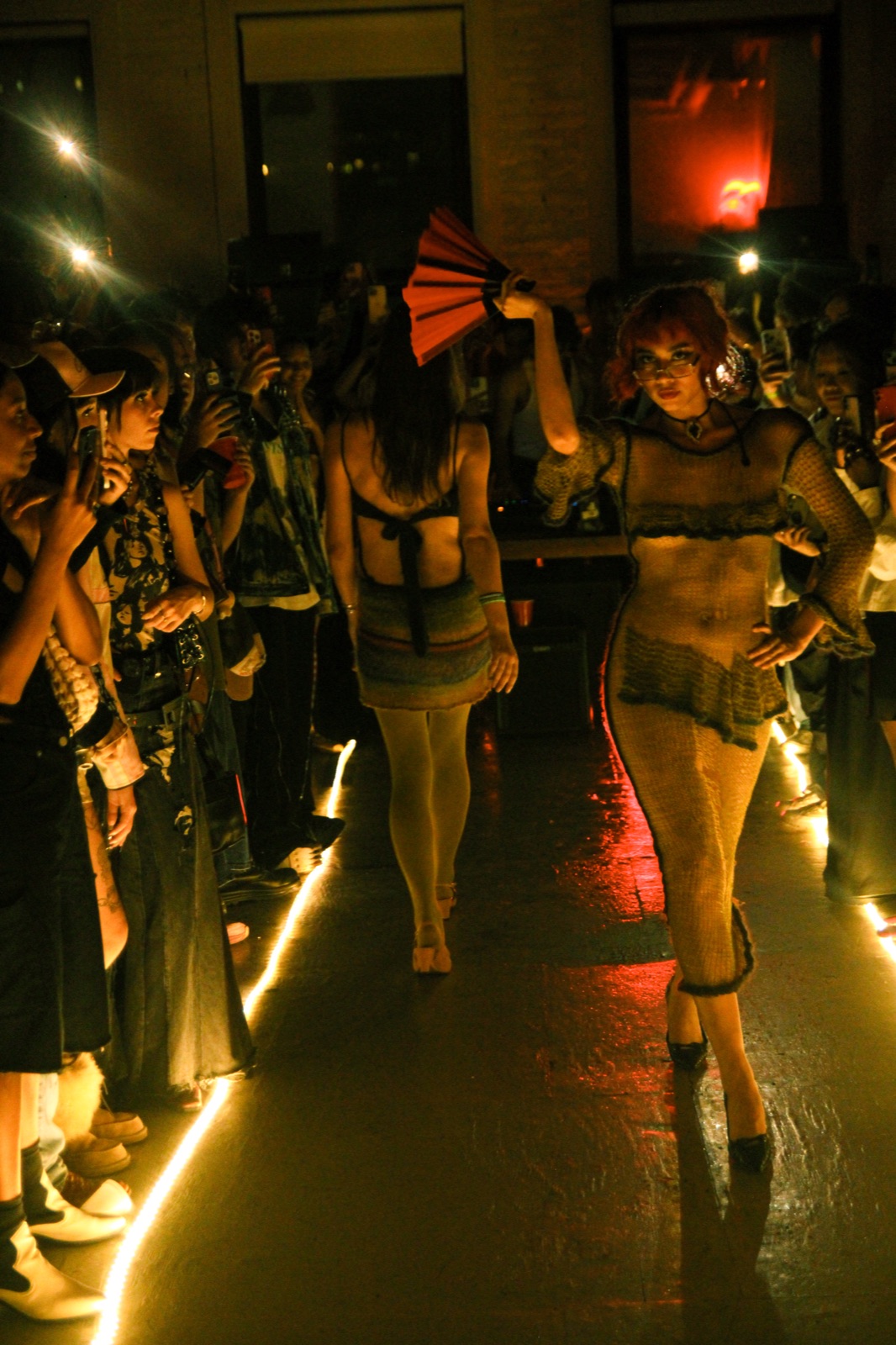 Models in dark looks walk the lit runway lane through a crowd in near darkness.