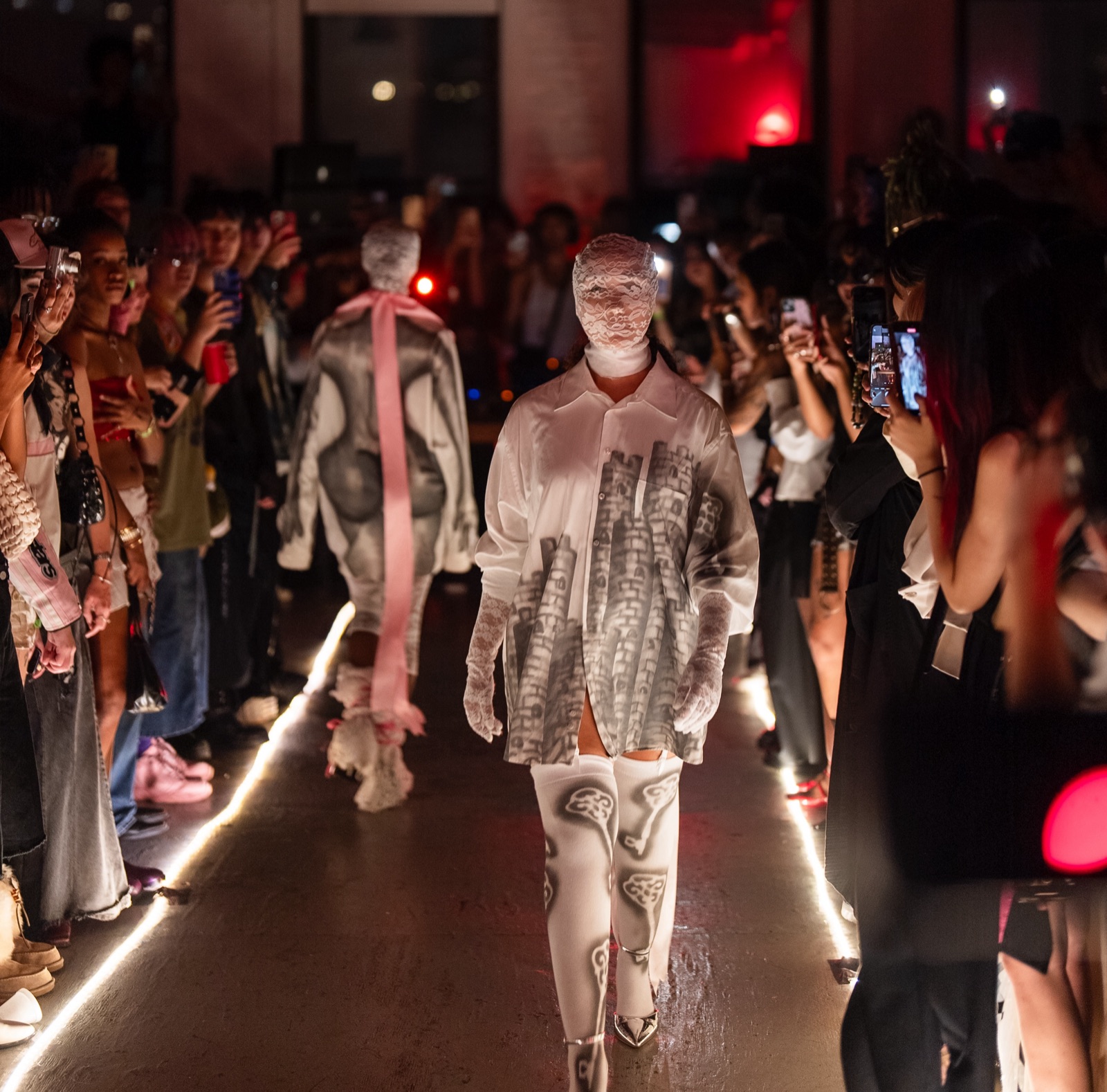 Models walk a runway outlined by glowing floor lights through a packed, dark warehouse crowd, phones raised.