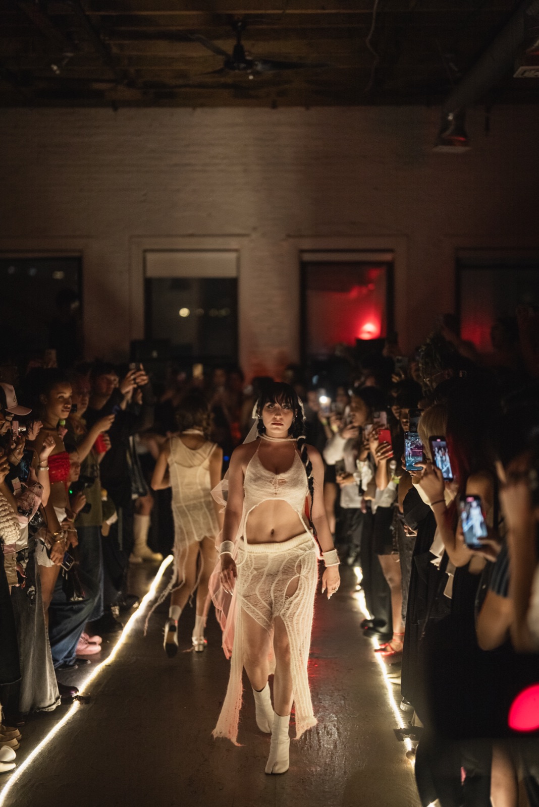 A model in a sheer cream outfit walks between two lines of floor lights as the crowd films.