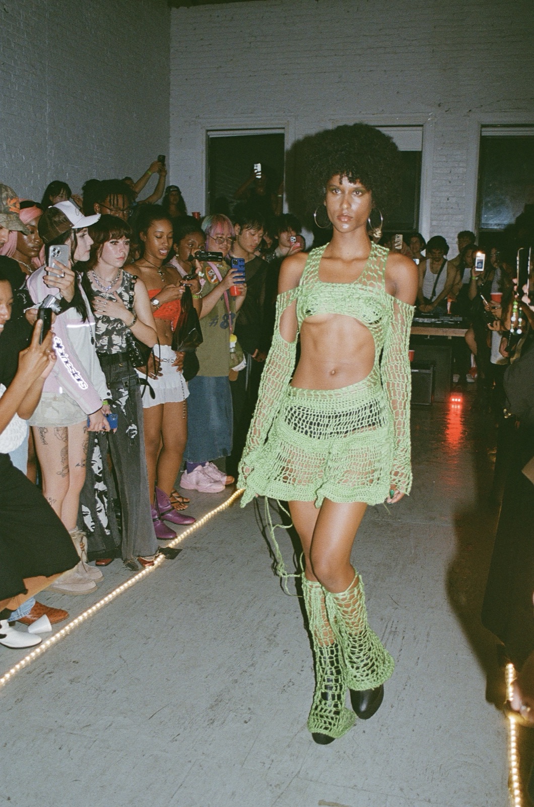 On film: a model in a green crochet set and lace-up boots walks past a line of onlookers.