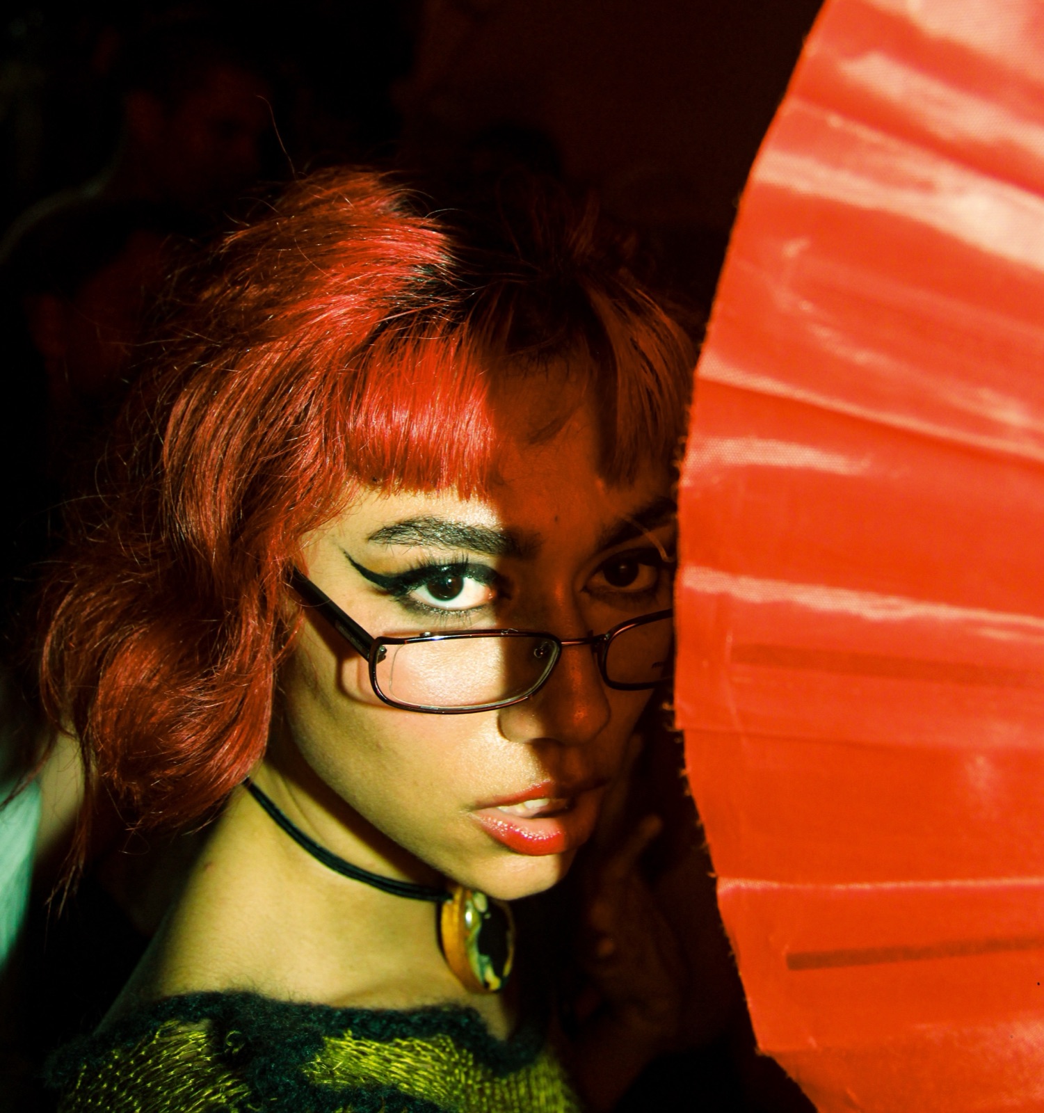 A person with red hair and glasses, half hidden behind a red folding fan.