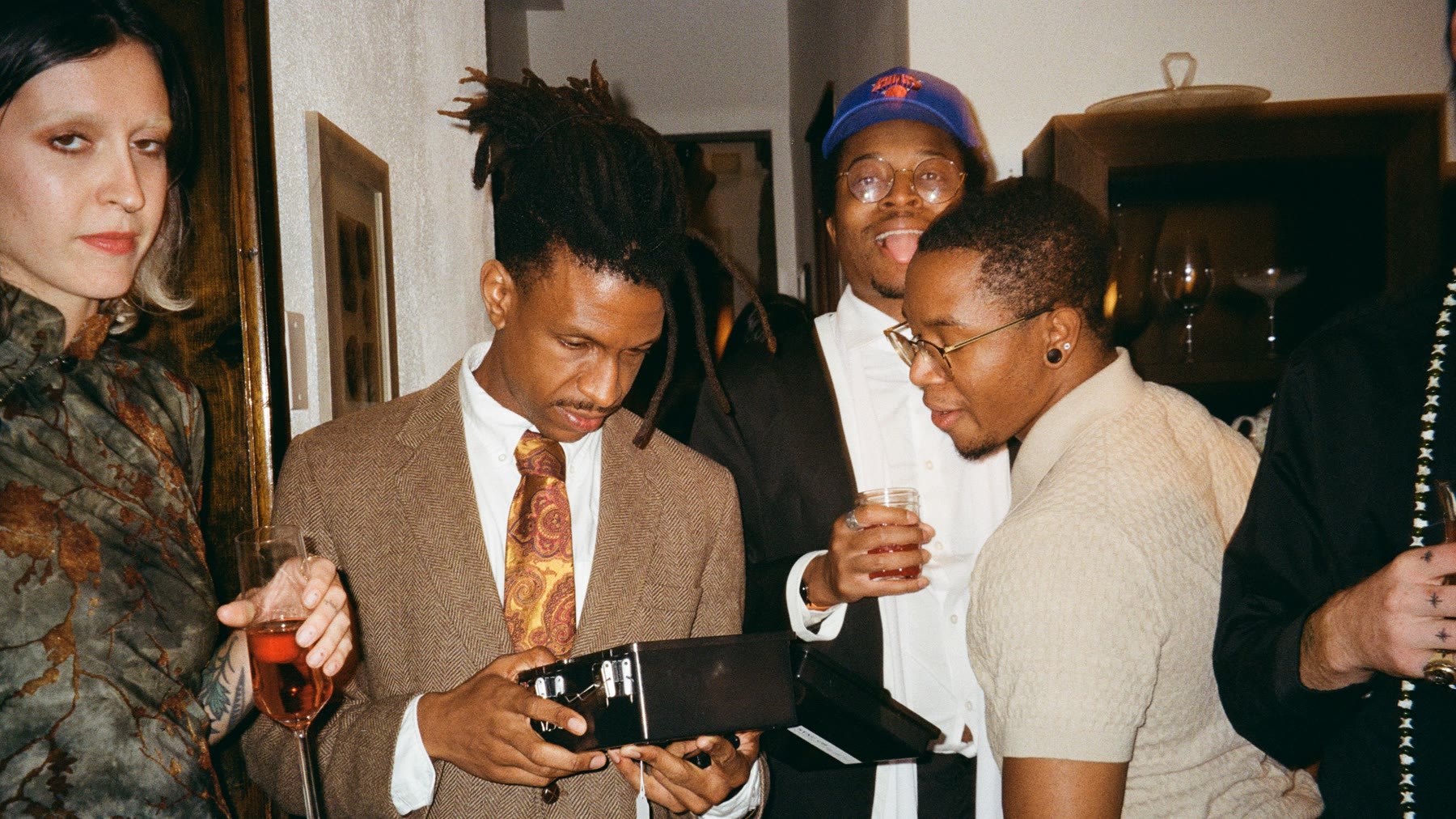 Guests in suits crowd around a player opening a black case in the castle hallway while a friend behind him sticks his tongue out at the camera.